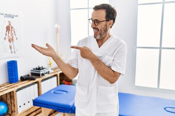 Middle age man with beard working at pain recovery clinic amazed and smiling to the camera while presenting with hand and pointing with finger.