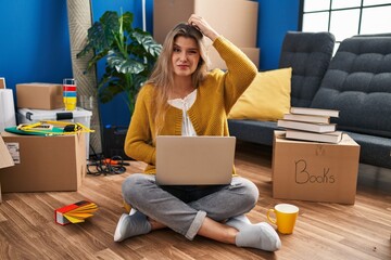 Young woman sitting on the floor at new home using laptop confuse and wonder about question. uncertain with doubt, thinking with hand on head. pensive concept.