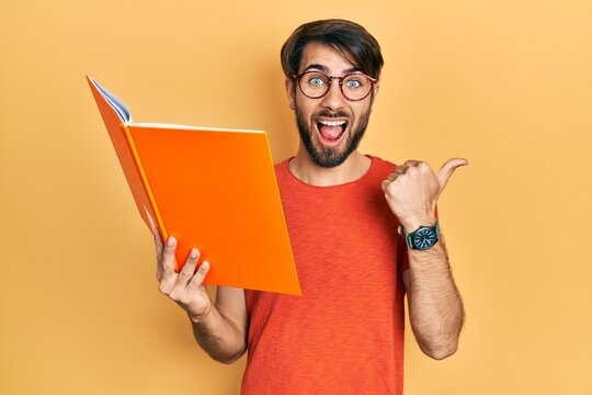 Young hispanic man reading book pointing thumb up to the side smiling happy with open mouth
