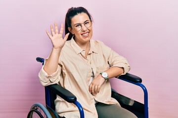 Young hispanic woman sitting on wheelchair waiving saying hello happy and smiling, friendly welcome gesture © Krakenimages.com