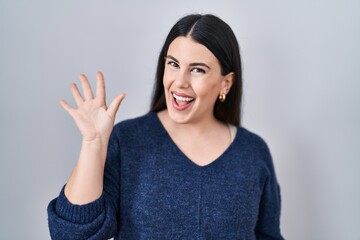 Young brunette woman standing over isolated background waiving saying hello happy and smiling, friendly welcome gesture
