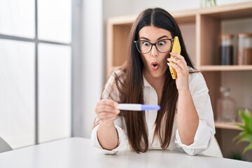 Young brunette woman holding pregnancy test result speaking on the phone afraid and shocked with...