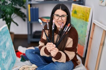 Young hispanic woman smiling confident holding paintbrushes at art studio