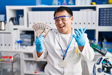 Young chinese man working at scientist laboratory holding money celebrating achievement with happy smile and winner expression with raised hand