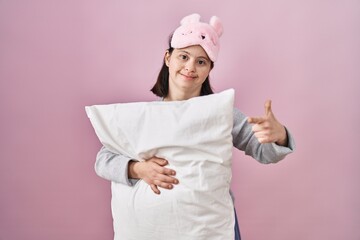 Woman with down syndrome wearing sleeping mask hugging pillow cheerful with a smile on face pointing with hand and finger up to the side with happy and natural expression