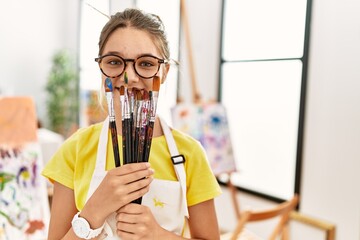 Adorable girl covering mouth with paintbrushes at art studio