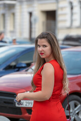 Naklejka na ściany i meble Young stylish woman wearing red dress and red lipstick walking on the city street near cars. Plus size model.