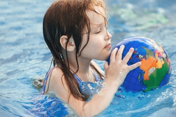 a little girl with wet hair, in a bright blue swimsuit in an outdoor pool in the summer plays a ball on the water