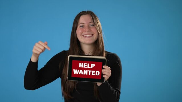 Smiling happy woman holding tablet computer with Help Wanted on screen on solid blue background.