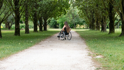 Fototapeta premium Young woman with long hair, moving in a wheelchair, from behind, in the middle of an alley lined with tall green trees