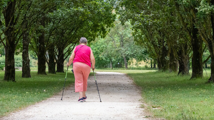 Woman in pink, from behind walking with sticks, on a leafy, green path