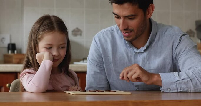 Happy engaged handsome dad and cute daughter kid moving pieces on board, playing learning game at kitchen table, thinking over tactic, talking, smiling, laughing. Fatherhood concept