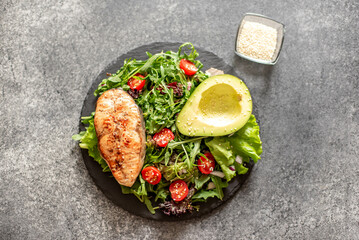 grilled salmon steak and fresh green vegetable tomato salad with lettuce and avocado on stone background
