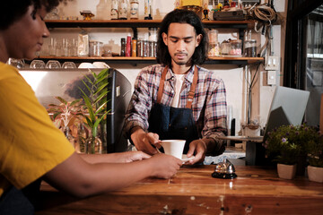 In front close-up, A young long hair Thai male cafe barista works by serving a cup of coffee to African American female customer at counter bar of coffee shop, happy service job, and SME entrepreneur.