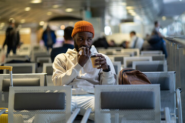 Young African American traveler man drinking coffee and eating sandwich while waiting for flying at...