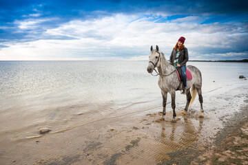 wearing jeans, jacket and autumn hat happy smiling female jokey goes on horseback along sea