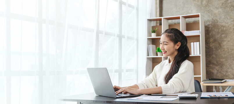 Happy confident young asian businesswoman sitting on her workplace in the office. Young woman working at laptop in the office.