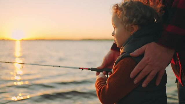 cute little boy is fishing by rod, father or grandpa is helping to child, happy family on river shore in sunset