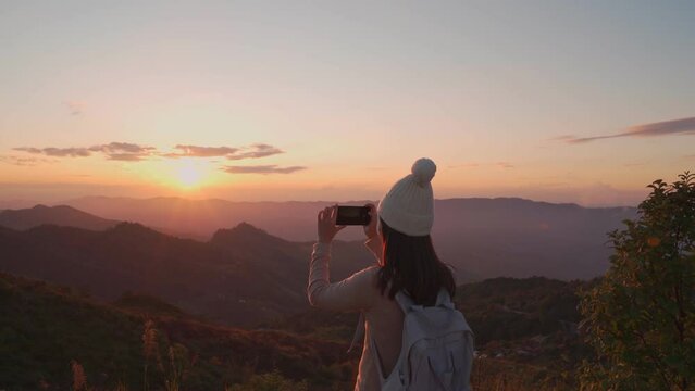 Happy young woman traveler taking photo with smart phone at the beautiful sunset on the top of the mountain