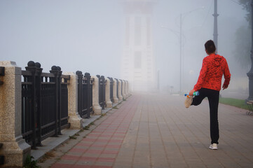 Girl athlete conducts training on a foggy embankment with a white lighthouse. View from the back. It stands on one leg. Water bottle in hand. Fencing of the promenade of their granite and metal. Blur.