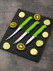a set of thin metal knives with a plastic handle on a black cutting board with chopped vegetables