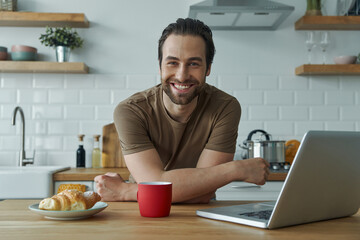 Fototapeta na wymiar Cheerful man enjoying breakfast and working on laptop while sitting at the kitchen island