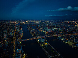 Aerial view of Queensboro Bridge and Roosevelt Island after sunset. Evening panoramic shot of city lights in urban boroughs. New York City, USA