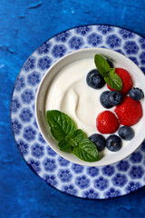 Natural yoghurt bowl close up photo. Sweet breakfast on blue stone table. Strawberry, blueberry, cherry, mint leaves on Greek yogurt. Eating healthy concept. 