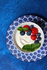 Natural yoghurt bowl close up photo. Sweet breakfast on blue stone table. Strawberry, blueberry, cherry, mint leaves on Greek yogurt. Eating healthy concept. 