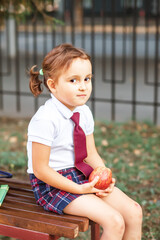 little cute schoolgirl in uniform sitting on a bench and eating an apple