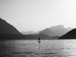 Grayscale shot of a person paddling on a board in the middle of a lake surrounded by lush greenery