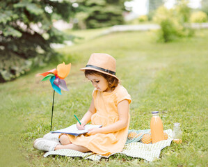 Cute little girl in a hat sitting on a blanket and writing or drawning at a picnic in nature