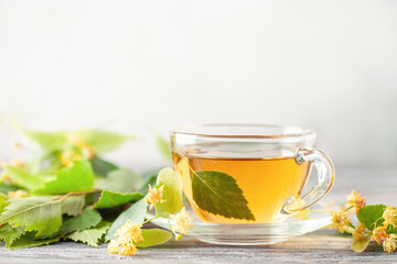 Glass teacup of green tea and linden flowers and birch leaves on wooden background. Herbal hot drink