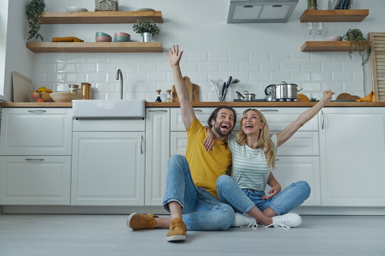 Cheerful couple embracing and keeping arms raised while sitting on the floor at the kitchen
