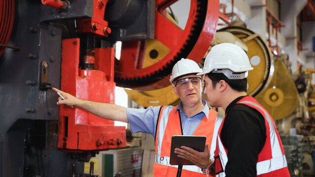 Senior man engineer wearing safety goggles and helmet standing holding tablet with coaching young engineer using hydraulic pump machine in factory.