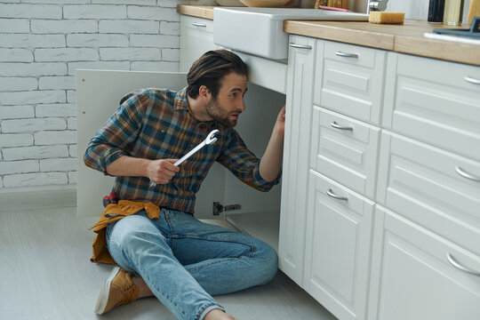 Confident young handyman using wrench while fixing a sink at the domestic kitchen