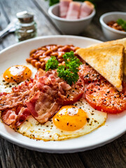 English breakfast - toasts, sunny side up eggs, bacon ,beans in sauce and fried tomato on wooden table
