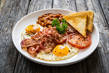 English breakfast - toasts, sunny side up eggs, bacon ,beans in sauce and fried tomato on wooden table
