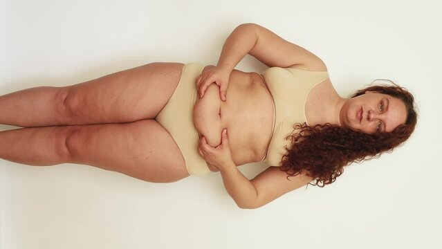 Vertical shot of red-haired woman in underwear looking at camera and her cellulite body. Squeezing large hydropic belly, hiding sag skin. Get rid of visceral fat. Fast gaining weight after childbirth