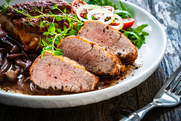 Roasted pork loin with shimeji mushrooms in sauce, fresh thyme, greens and cherry tomatoes on wooden table
