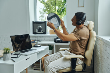 Confident African man in headphones examining vinyl record while sitting at working place