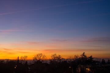 Colourful sunset with clear sky and some clouds from the city, with the silhouettes of trees and some buildings in front of the mountains on the horizon