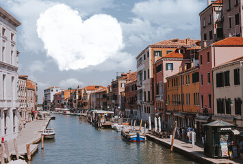 Fototapeta na wymiar Cloud in the shape of a heart in the sky over the city. Fabulous cityscape overlooking the canal with boats and people walking the streets of Venice. A wonderful tourist day.