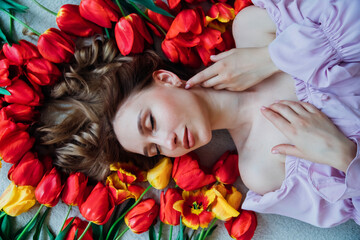 Obraz na płótnie Canvas A young woman is lying on the floor among red tulips. The concept of March 8, Valentine's Day. Spring portrait of a woman.