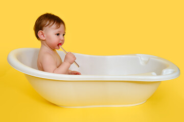 Happy baby toddler boy brushes his teeth with a toothbrush in a white tub on a studio yellow...