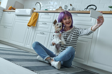 Happy woman carrying little dog and making selfie while sitting on the floor at the kitchen