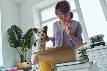 Beautiful young woman unpacking box while her cute dog sitting near her on the table
