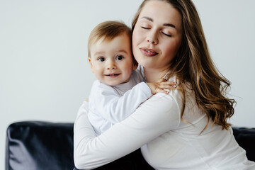 Young woman cuddling talking to small kid son, having fun together on couch in studio living room. Smiling child boy communicating with elder sister nanny babysitter, laughing at home.
