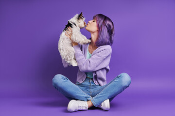 Obraz na płótnie Canvas Playful young woman carrying little dog while sitting against purple background