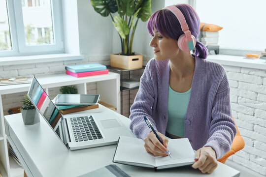 Confident young woman in headphones making notes while sitting at her working place